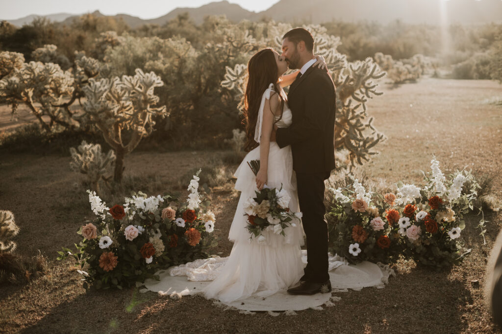 Photo of a bride and groom in the Arizona desert wearing a wedding dress and black suite surrounded by flowers and cactus.