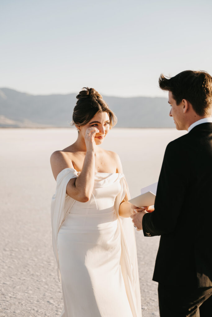 Untraditional wedding at the Utah salt flats with a bride in a wedding dress and groom in a black suit reading private vows to each other.