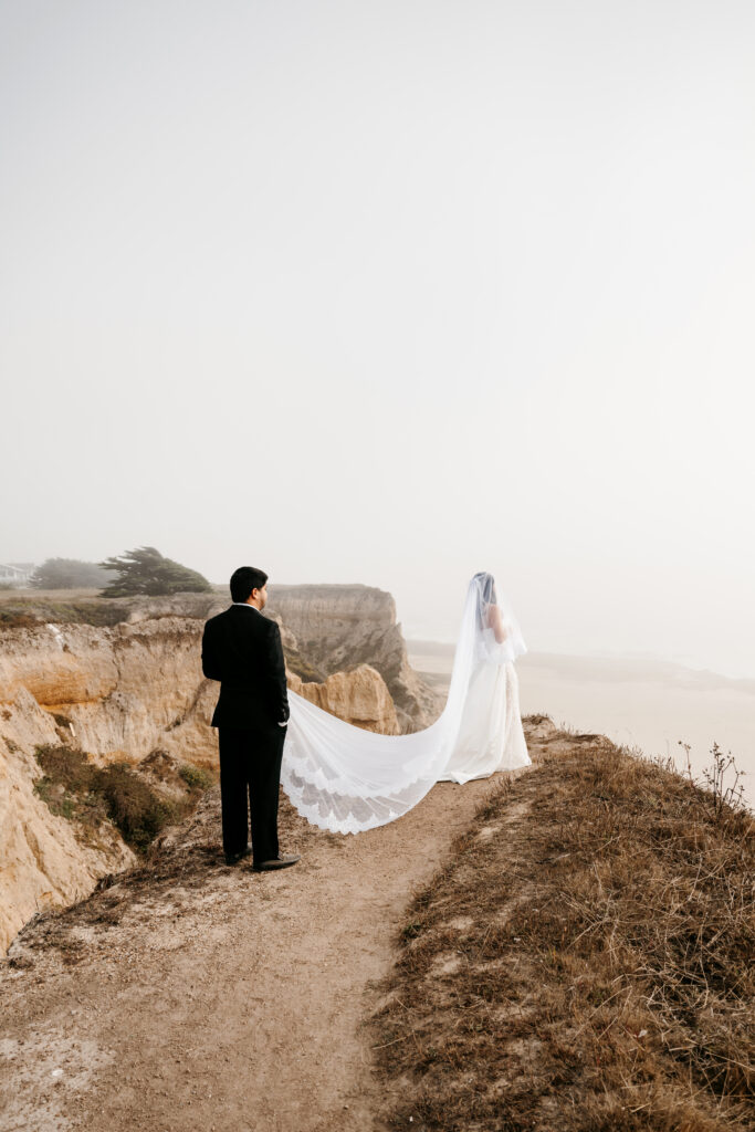 Bride and groom standing on a cliff side looking out at the ocean in California