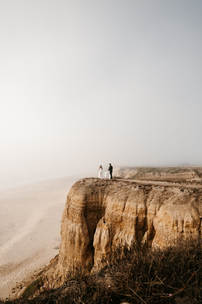Bride and groom standing on a cliff side looking out at the ocean in California